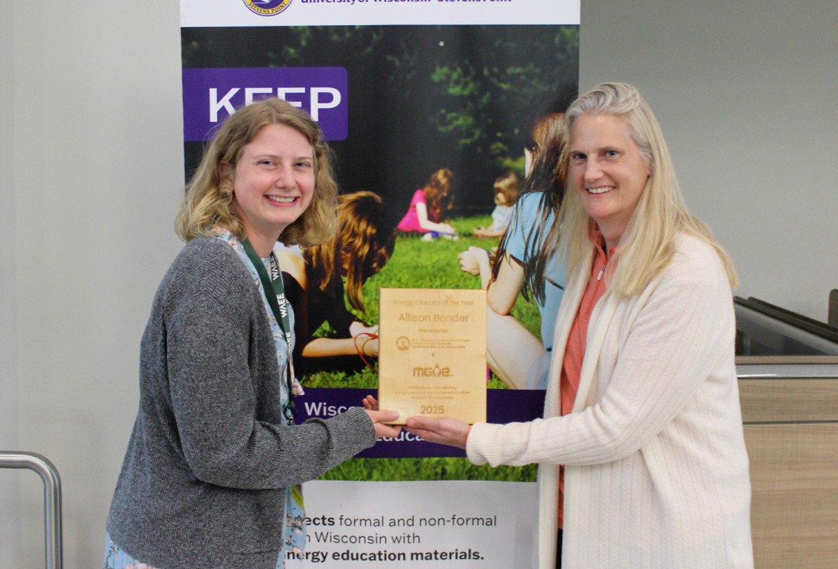Two women smiling and holding a wooden plaque stand in front of a poster labeled KEEP with a photo of children on green grass