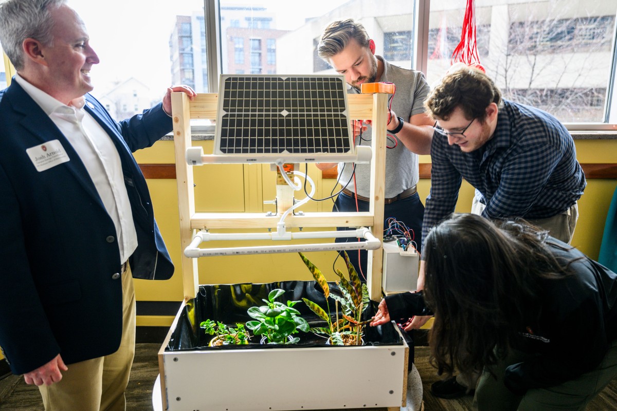 Four people look at a solar panel mounted atop a box containing plants.