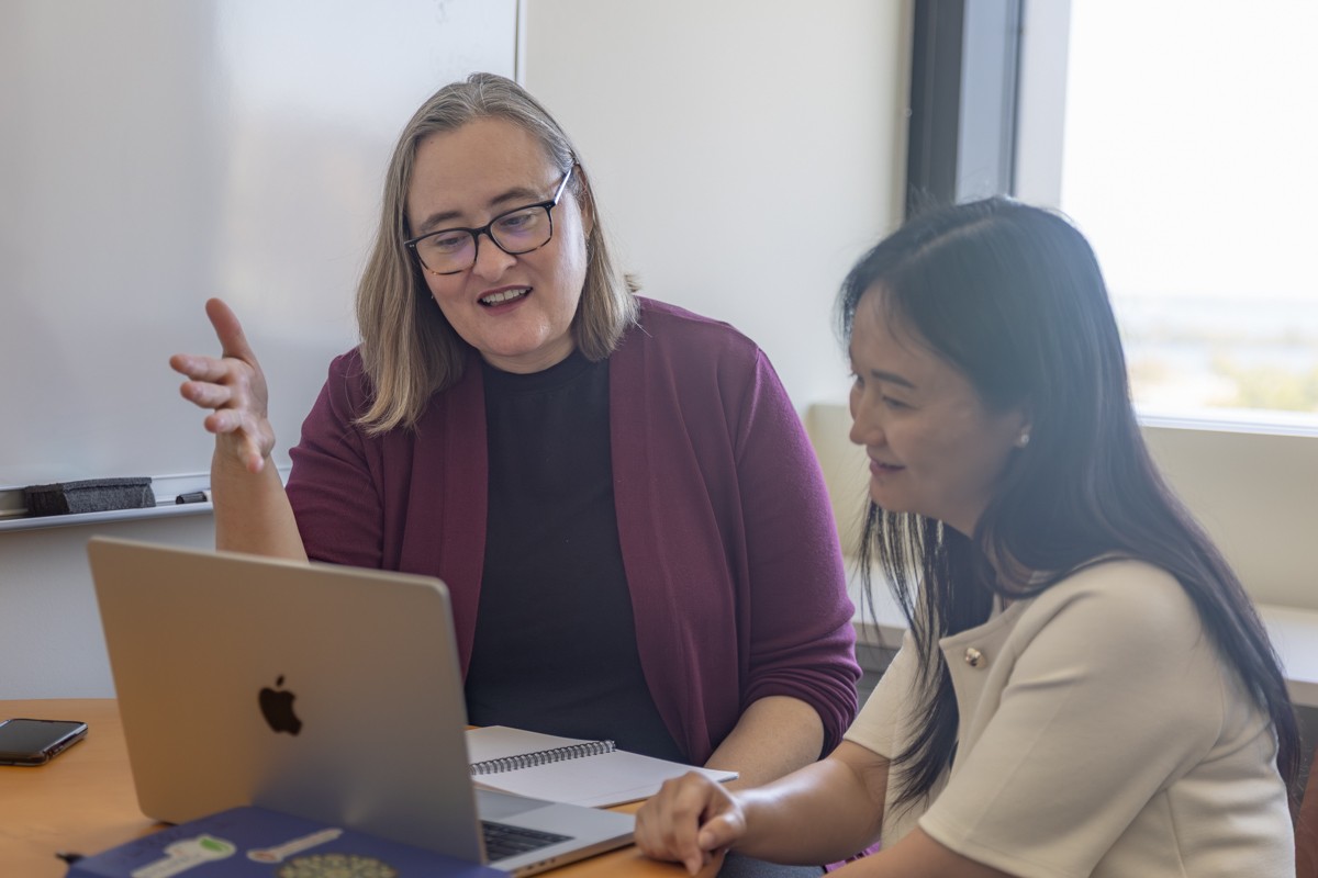 A photo of two women seated at a table and looking at a laptop screen. The woman on the left is gesturing with her right hand while the woman on the right looks on.
