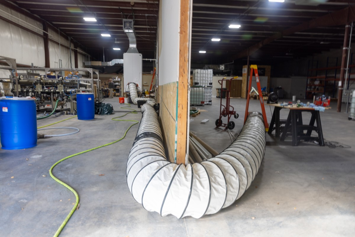 A large, mostly empty industrial space bisected by a wall. A flexible hose extends from the right side of the space to a machine on the left side, bending around the end of the wall.