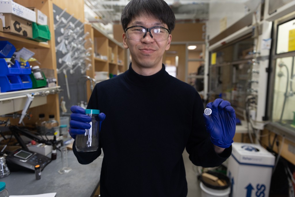 Smiling man standing in a laboratory holding a jar of black powder in one hand and a silver coin battery in the other