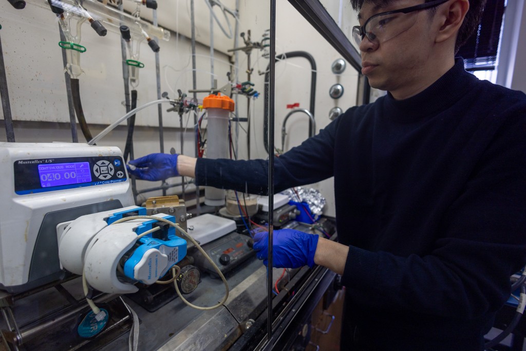 A researcher wearing blue nitrile gloves adjusts settings on a peristaltic pump connected to tubing and an electrochemical flow system inside a laboratory fume hood.