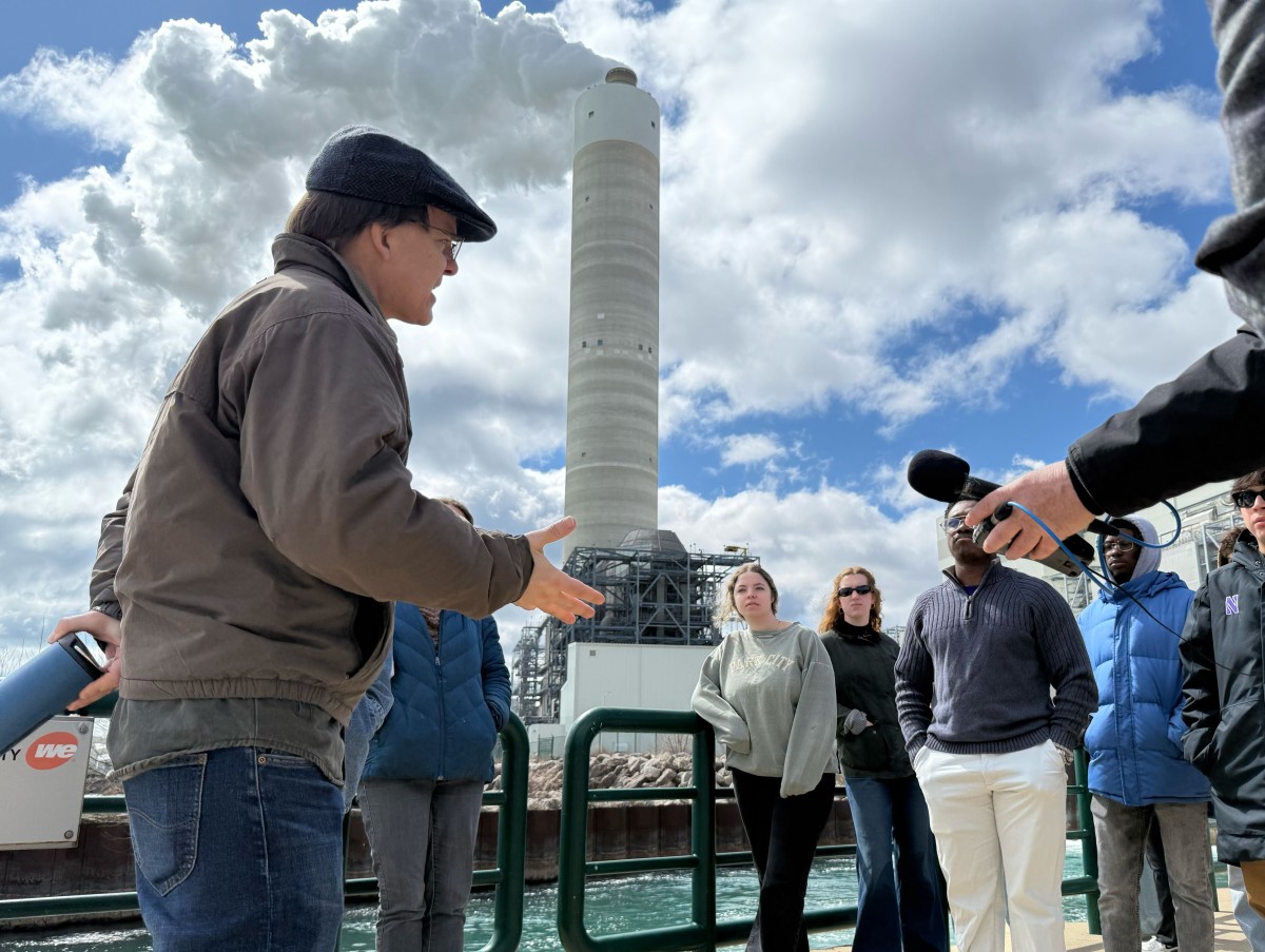 A man speaks to a group of people in front of a tall smokestack billowing white steam into a blue sky pocked with white clouds
