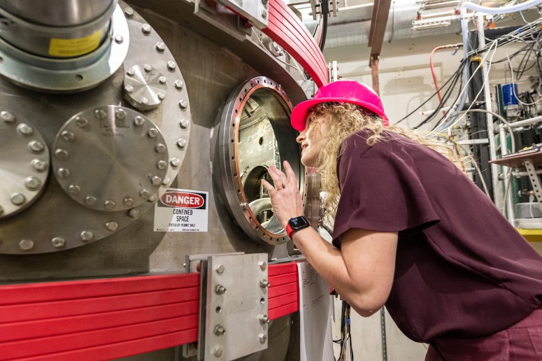 Woman wearing red hard hat peers through glass portal on a large, complex piece of scientific equipment