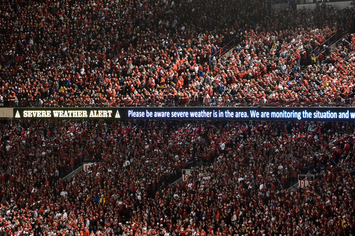 A densely packed stadium crowd, mostly dressed in red and white, watches a football game. A digital ribbon board across the stands displays a severe weather alert warning that severe weather is in the area.