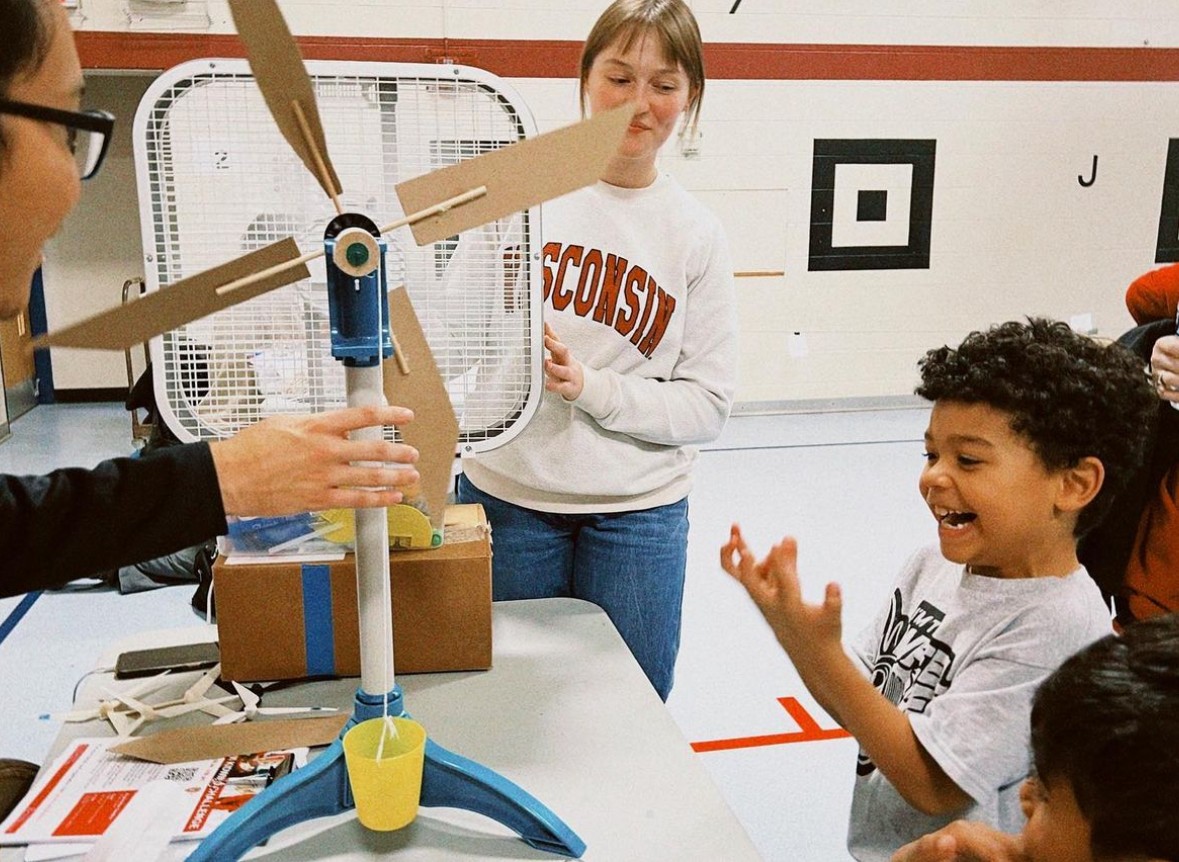 Child reacts with excitement as two team members demonstrate a model wind turbine in front of a box fan