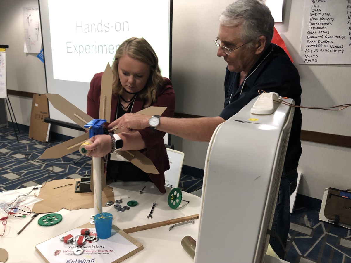 A teacher and the workshop instructor work on attaching a hub and blade set to the turbine in front of a box fan.