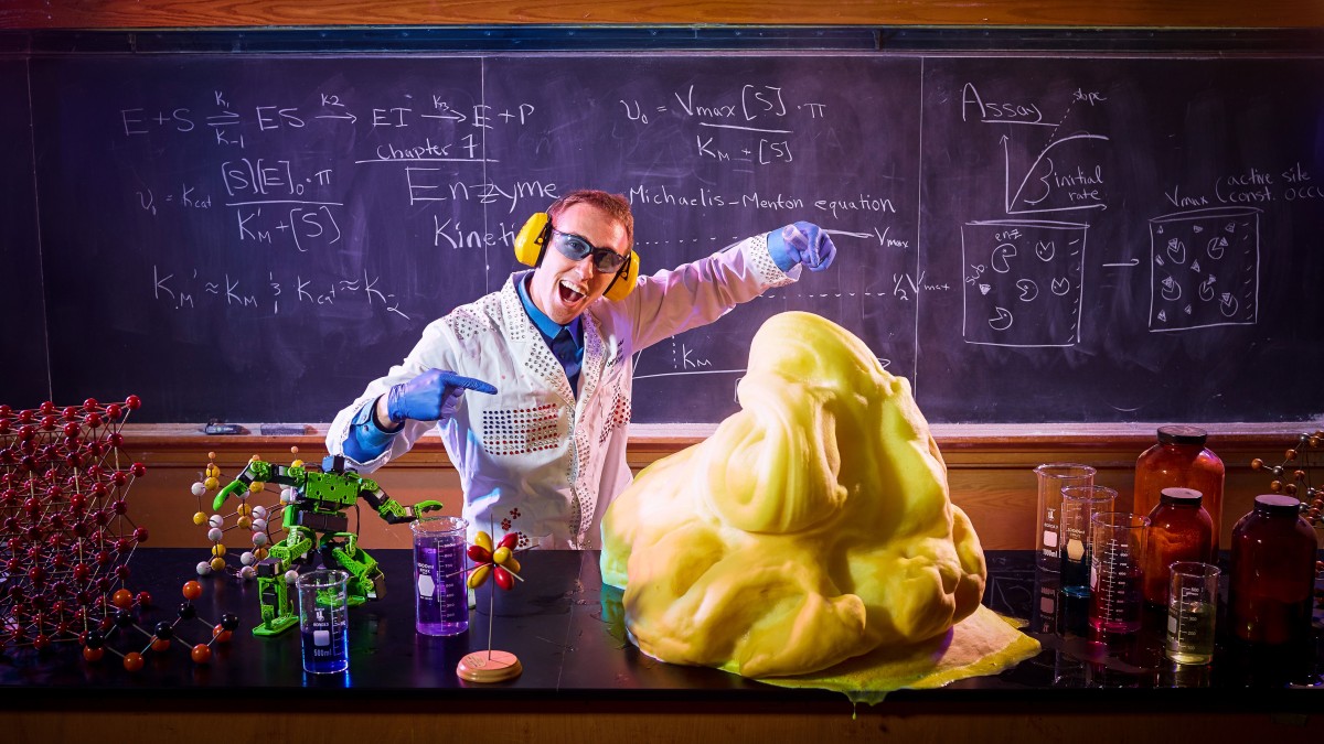 A man in a sequin-covered lab coat and blue rubber gloves stands in front of a chalk board covered with equations gestures at a large pile of yellow slime on a lab bench covered with beakers and models of molecules