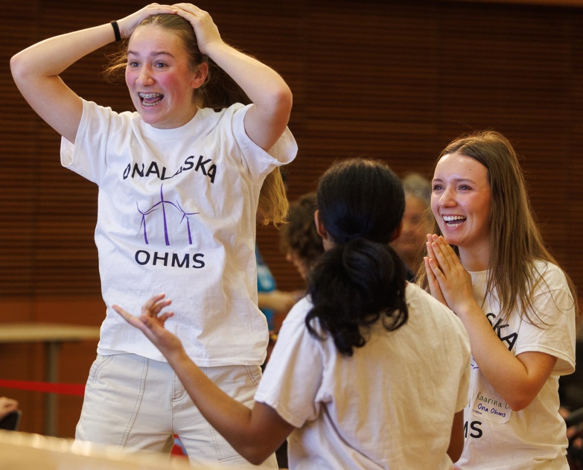 Three students wearing white "Onalaska Ohms" t-shirts react with extreme excitement. One girl has her hands on her head in joyous shock, while another smiles broadly with her hands clasped together.