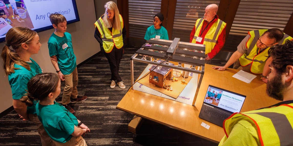 Four students in teal t-shirts stand around a table presenting their model solar home to four adult judges wearing bright yellow safety vests. A laptop on the table displays a presentation slide asking, "Any Questions?"