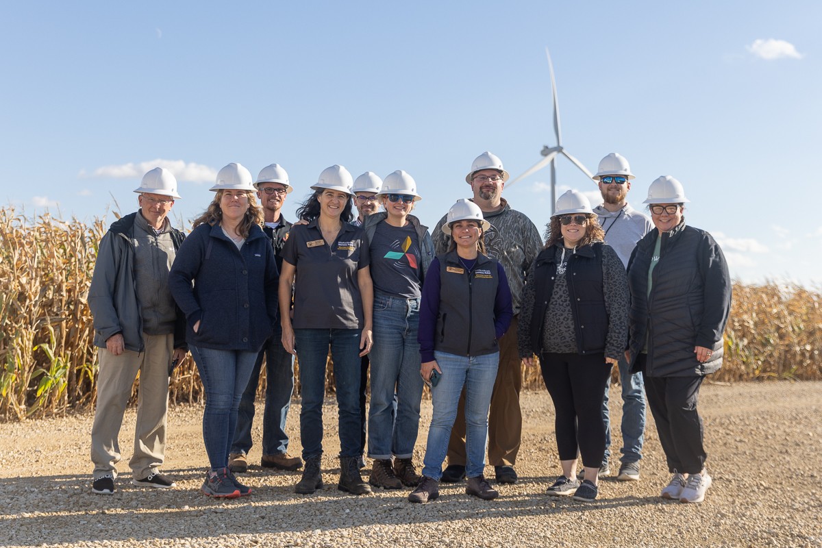 A group of teachers stands smiling in front of a corn field with a wind turbine in the distance.
