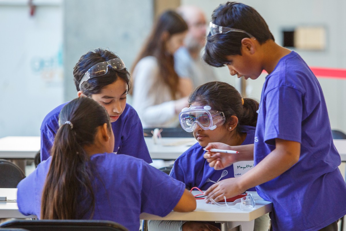 Four kids in purple T-shirts with safety goggles work at a table