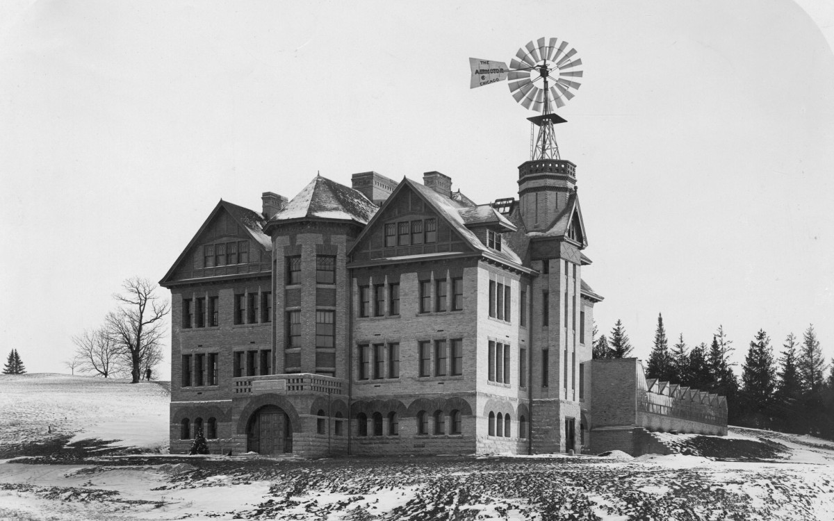 Black and white photo of a four-story stone building sitting alone on a hilltop. A windmill sits atop a turet on the right side of the building.