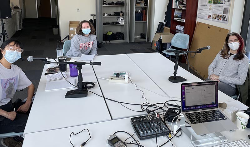 Michelle Chung, left, sits at a big table while interviewing guest Dominique Brossard, right. Mary Riker takes notes and prepares questions with a notebook, center.