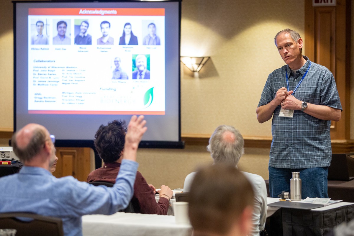 A researcher presents at a scientific conference, fielding a question from an audience member with a raised hand, with an acknowledgments slide listing collaborators and funders projected on the screen behind him.