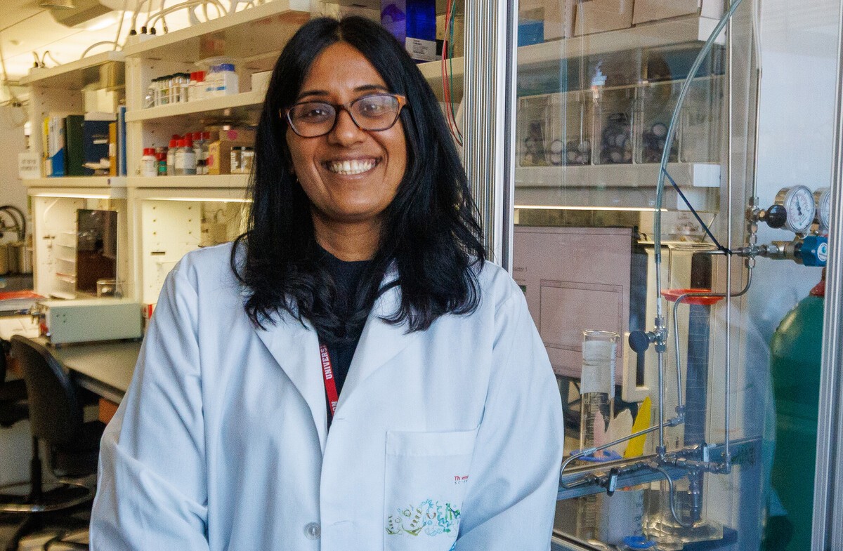 A smiling researcher in a white lab coat and glasses stands in front of a fume hood and chemical shelving in a chemistry laboratory.