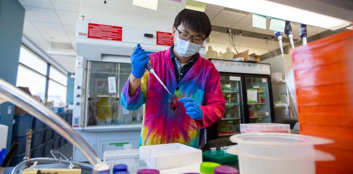 Sae-Byuk Lee stands in front of a lab bench pipetting samples in his tie-dyed lab coat