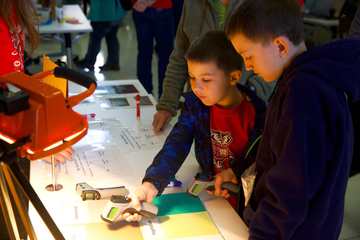 Two young boys, with faces lit in soft lamp light, hold thermometers to sheets of paper.