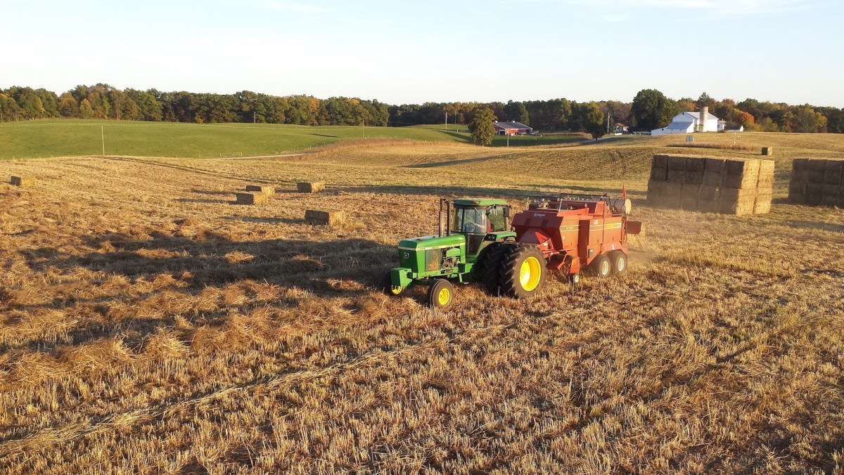 A green tractor pulls a red baler in a field of brown grass. Rectangular bales of grass are stacked in the background and scattered through the field. A line of grass and trees with a white barn are visible in the background.