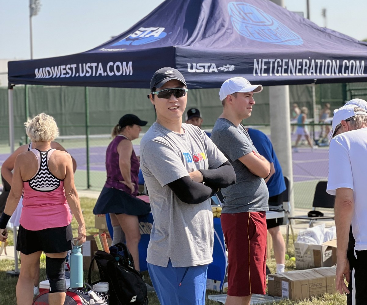 Man in sunglasses, baseball cap, and T-shirt stands in front of canopy with the words "midwest.usta.com"