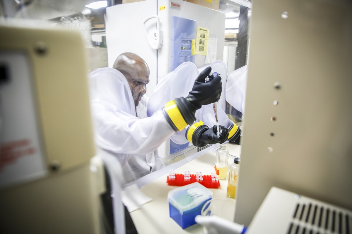 Victor Ujor pipetting a yellow liquid from a bottle inside an enclosed Plexiglass chamber