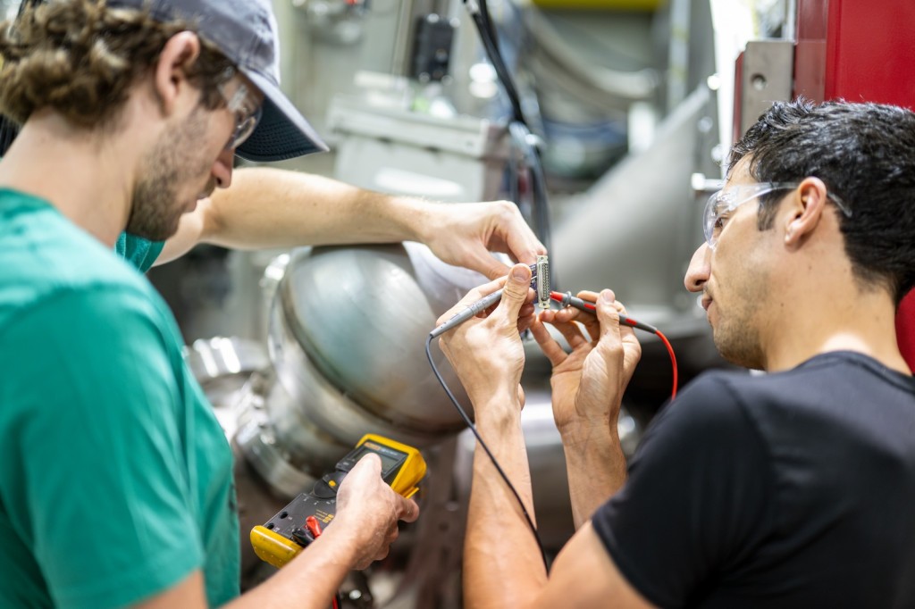 Two men work on equipment in a lab setting. The man on the right holds two wire probes to a piece of metal as the one on the left holds a metering device.