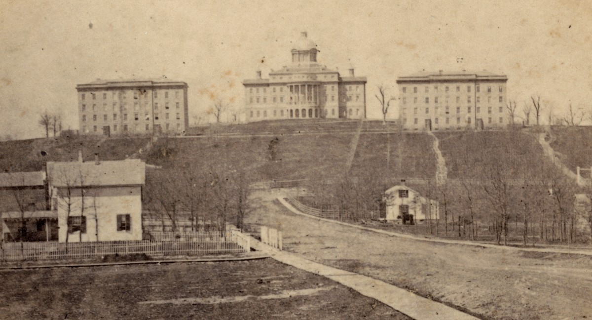 Sepia-tinted photo showing three large buildings atop a hill. In the foreground are two wooden homes on either side of a dirt road.