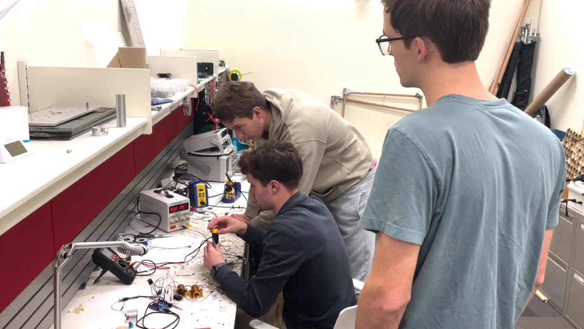 Three students work together at an electronics workbench, with one soldering a circuit while the others observe, surrounded by a power supply, soldering station, multimeter, and various wires and components.