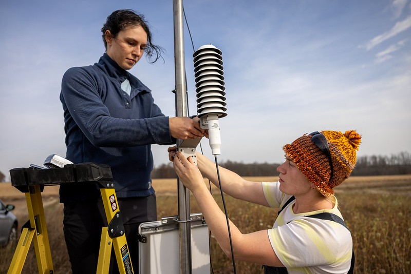 Two researchers in an open field work together to mount a white, louvered weather sensor onto a metal pole, one standing beside a yellow ladder and the other, wearing an orange knit hat, reaching up to secure the instrument.