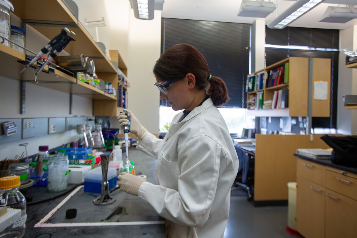 A woman in a lab coat and safety glasses uses a pipette at a lab bench cluttered with jars, tubes, and supplies, with bookshelves visible behind her.