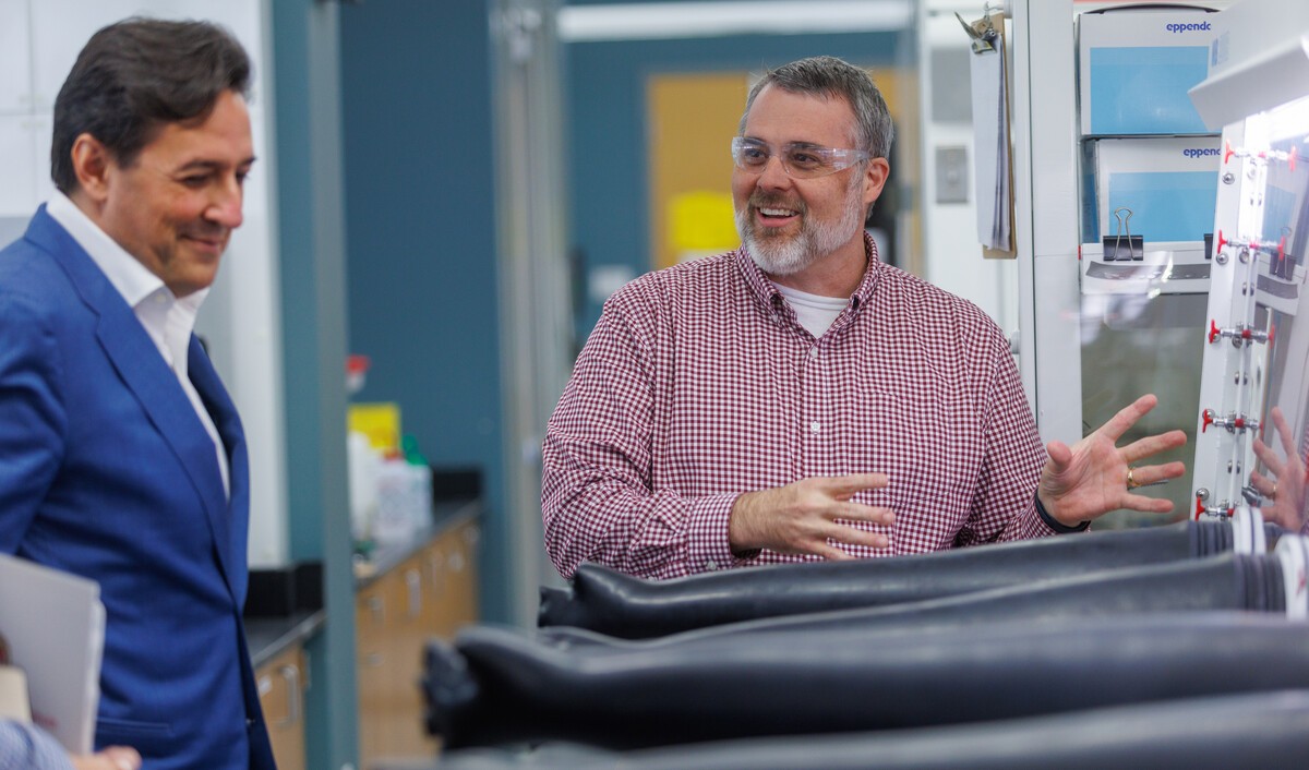 A man in a blue blazer looks on with a smile as a bearded man in a red checked shirt and safety glasses gestures animatedly beside a glovebox with black gloves in a laboratory.
