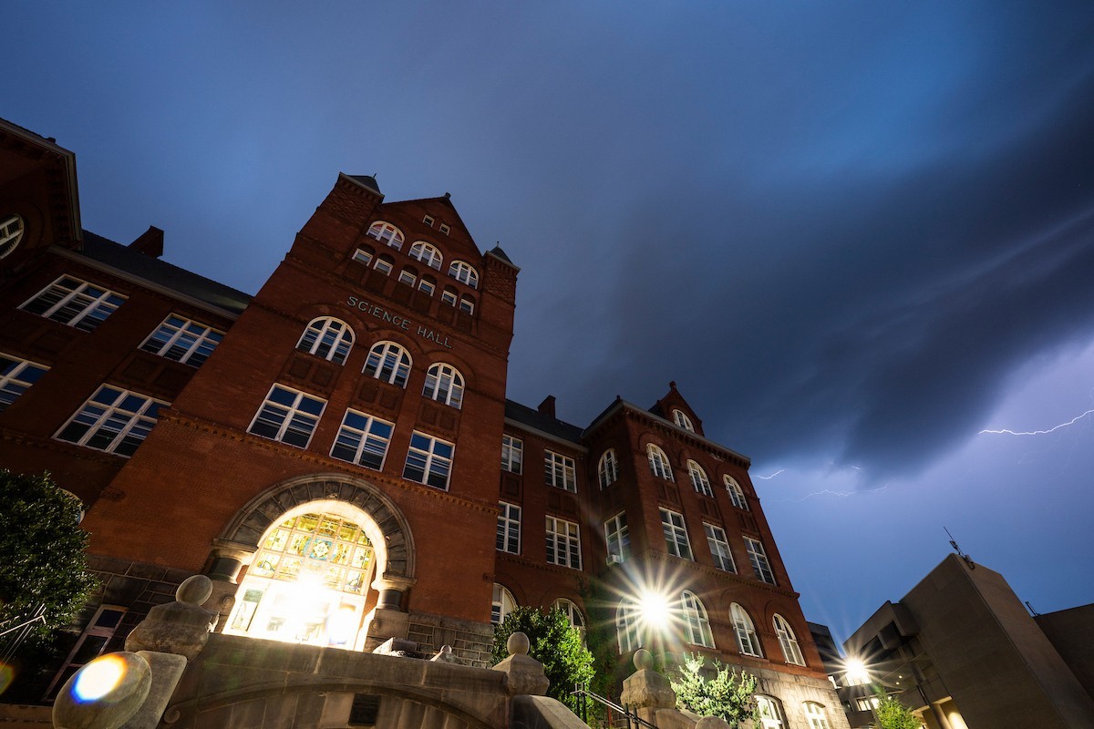 A red-brick university building labeled “Science Hall” is illuminated at night beneath dark storm clouds, with a bright lightning bolt visible in the sky to the right.