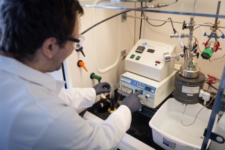 Man in white lab coat reaches into an exhaust hood to adjust equipment