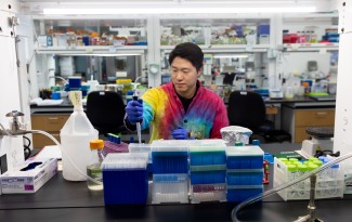 Asian man in tie-died lab coat seated at a lab bench