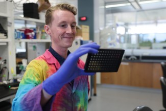 A blonde man in a the-dyed lab coat holds a square tray in one blue gloved hand