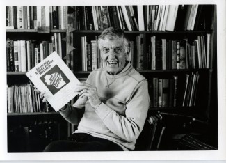 black and white photo of smiling man seated in front of a bookshelf holding a bound paper titled "Alternative energy futures for Wisconsin"