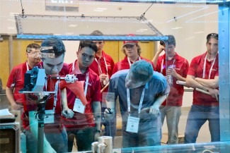 A man in a blue shirt leans in to observe as man in red shirt adjusts mechanism on a model wind turbine while a group of other men in red shirts look on