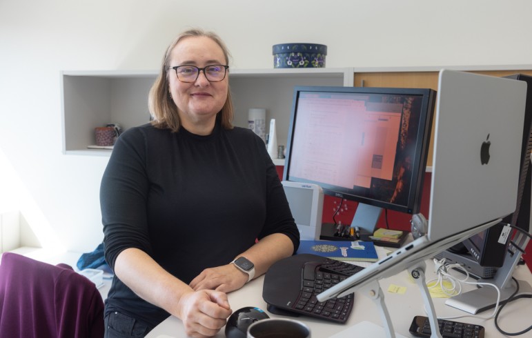 Woman with blonde hair and glasses sits at a desk with a computer monitor. She is turned to her right to face the camera