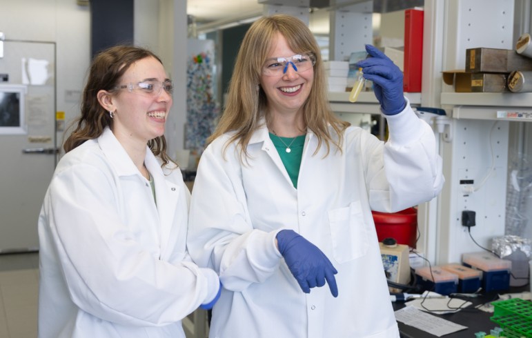 A woman in a white lab coat and safety glasses holds up a flask of opaque yellow liquid as another woman in similar attire looks on smiling