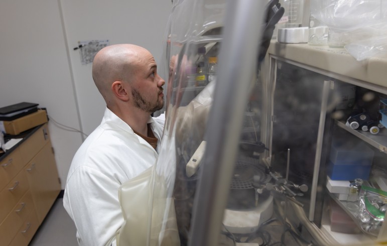 A bearded man in a white lab coat reaches for an object as he stands in front of a clear plastic barrier