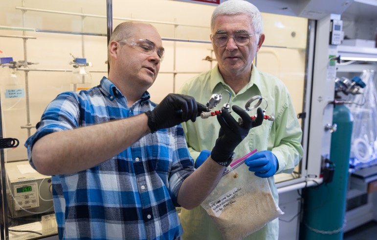 Man wearing black rubber gloves holding a piece of lab equipment as man holding plastic bag of wood chips looks on