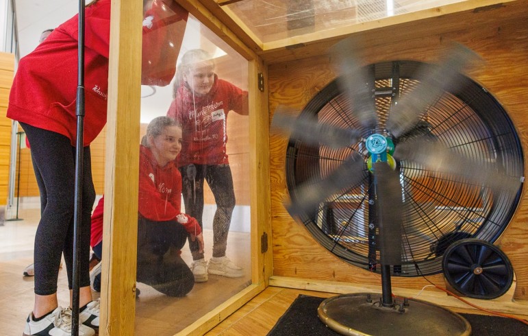 Three girls wearing matching red "Wind Storm" hoodies look through a clear panel into a wooden wind tunnel, watching a large fan spin rapidly.