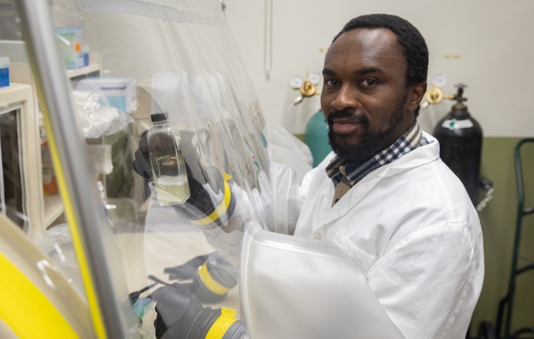 A man in a white lab coat holds a tube full of opaque liquid under a plastic sheet