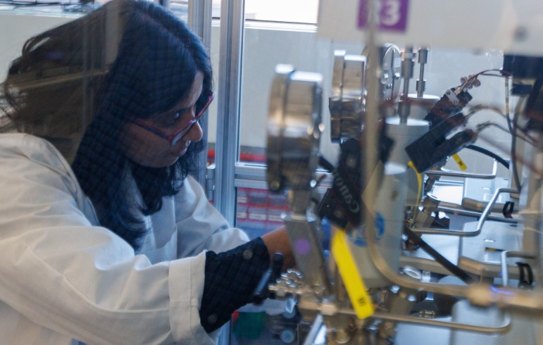 A researcher in a lab coat, safety glasses, and nitrile gloves sets up a high-pressure reactor system in a glass enclosure.