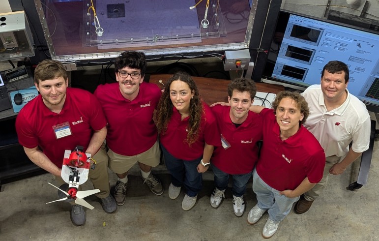 The WiscWind student team in matching red polos poses in front of a wind tunnel and data acquisition monitors, with one member holding their small-scale wind turbine prototype.