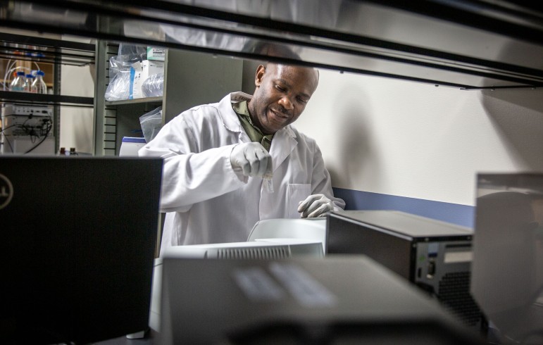 Man in white lab coat framed by dark shelving holds a glass vial