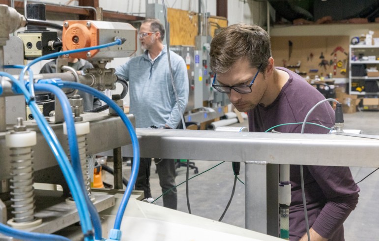 A man peers down at a piece of machinery with coil springs and blue tubingin an industrial setting. In the background a second man looks at something off camera.