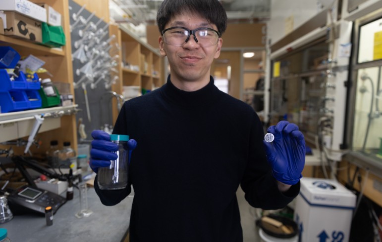 Smiling man standing in a laboratory holding a jar of black powder in one hand and a silver coin battery in the other
