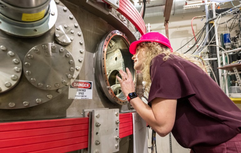 Woman wearing red hard hat peers through glass portal on a large, complex piece of scientific equipment