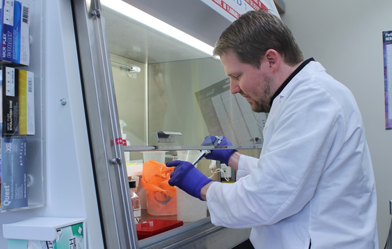 A man in a white lab coat and blue latex gloves pipettes liquid under a ventilation hood.